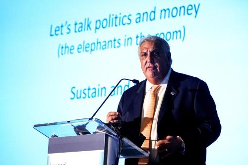 Person speaking at a podium at the Beyond Flexner conference. Presentation behind reads "Lets talk politics and money (the elephant's in the room)"