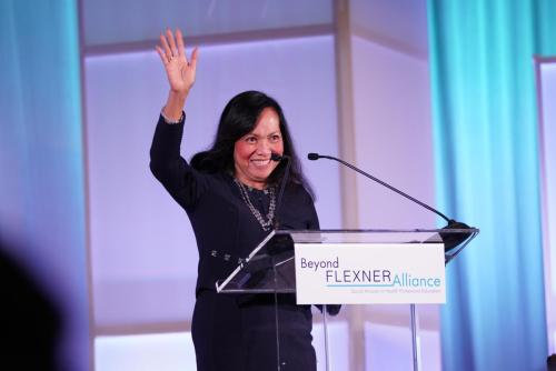 Woman waving at the crowd from a podium with a sign that says Beyond Flexner